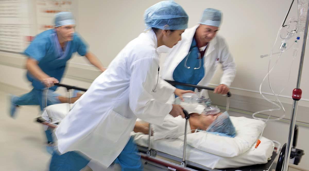 Three hospital workers in scrubs manuever a patient on a gurney through a hallway in a medical facility.