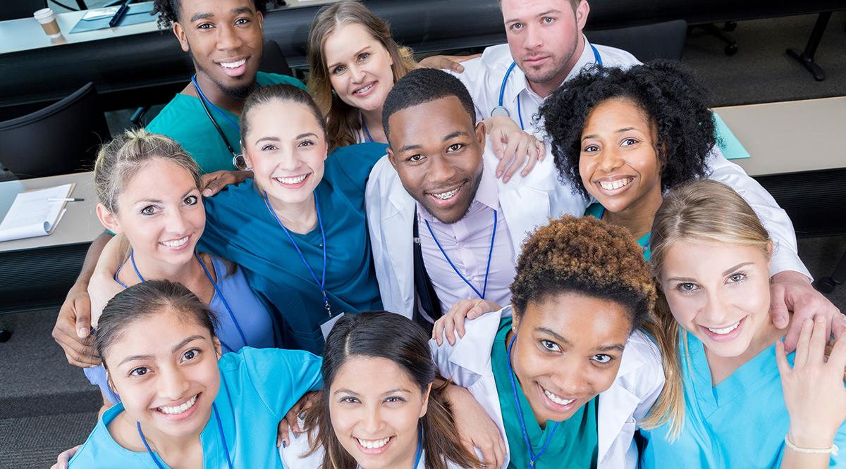 This is a view from above of a group of medical students looking up and smiling for the camera. They are standing in a lecture hall with table behind them.