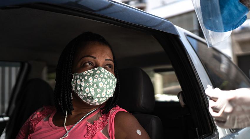 A masked woman in a car prepares to receive a vaccine from a masked worker