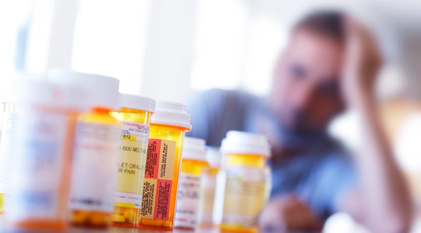 A large group of prescription medication bottles sit on a table in front of a distraught man.