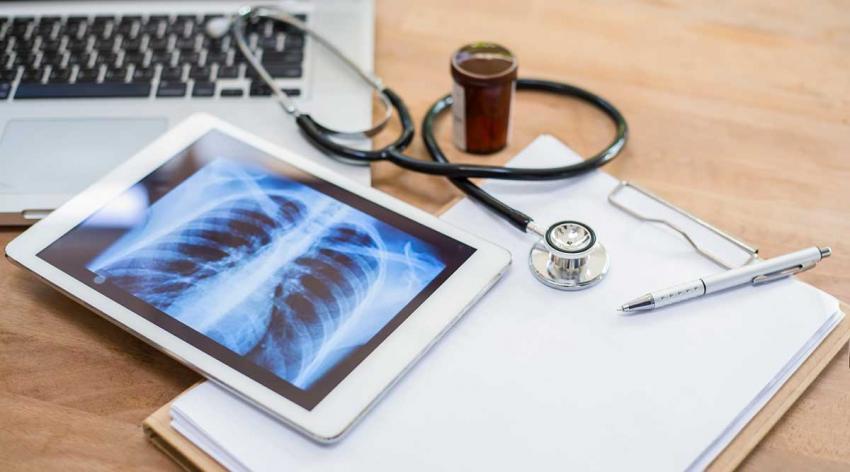 A laptop, a tablet showing a lung x-ray scan, a stethoscope, a notepad, and a pen on a table
