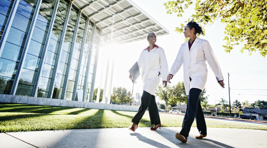 Two doctors in white coats walk outside on a school campus