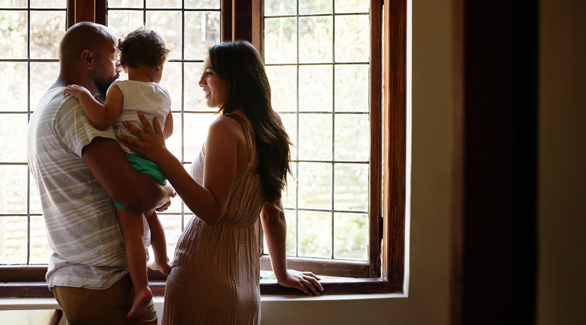 Two parents stand in front of a window holding a baby