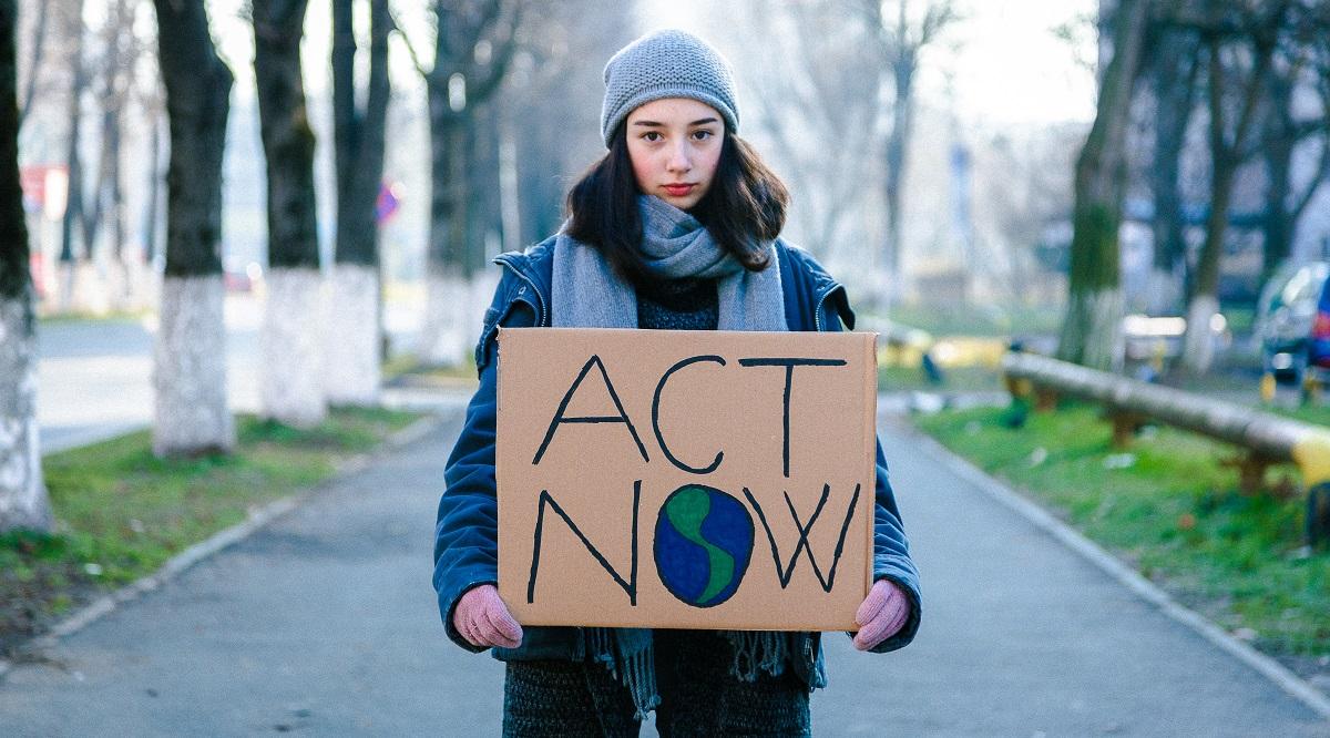 Young activist holding act now sign Portrait of a young teenage girl activist holding a sign protesting against climate change and global warming. She is standing outdoors on a city street and is holding signs with various messages, such as 'go green' and 'save our planet'. She is looking directly at the camera with a serious expression. Focus on the girl and her sign, with city street defocused beyond. Room for copy space.