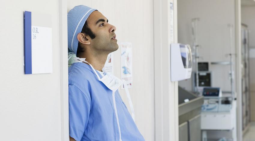 A doctor in scrubs stands against a wall looking tired