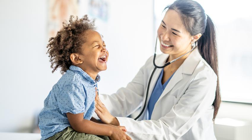 A sweet young mixed race boy, sits up on an exam able with a big smile on his face, during a routine check-up. His female doctor of Asian decent is leaning in with her stethoscope to listen to his heart.