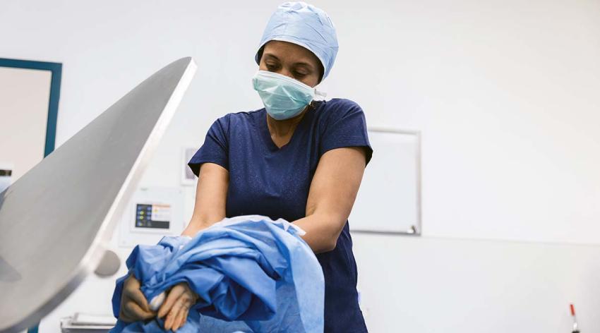 A female doctor wearing blue scrubs, a surgical mask, and a cap, removes her protective gown.