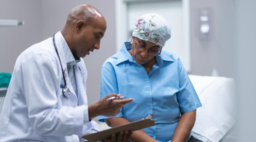 Patient and doctor are sitting next to each other on an examination table in a medical clinic. The doctor is asking the patient questions and taking notes on a clipboard.